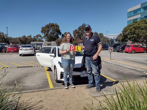 A learner driver holding a P plate standing next to a volunteer L2P mentor holding an L plate. They both stand in front of the L2P car.