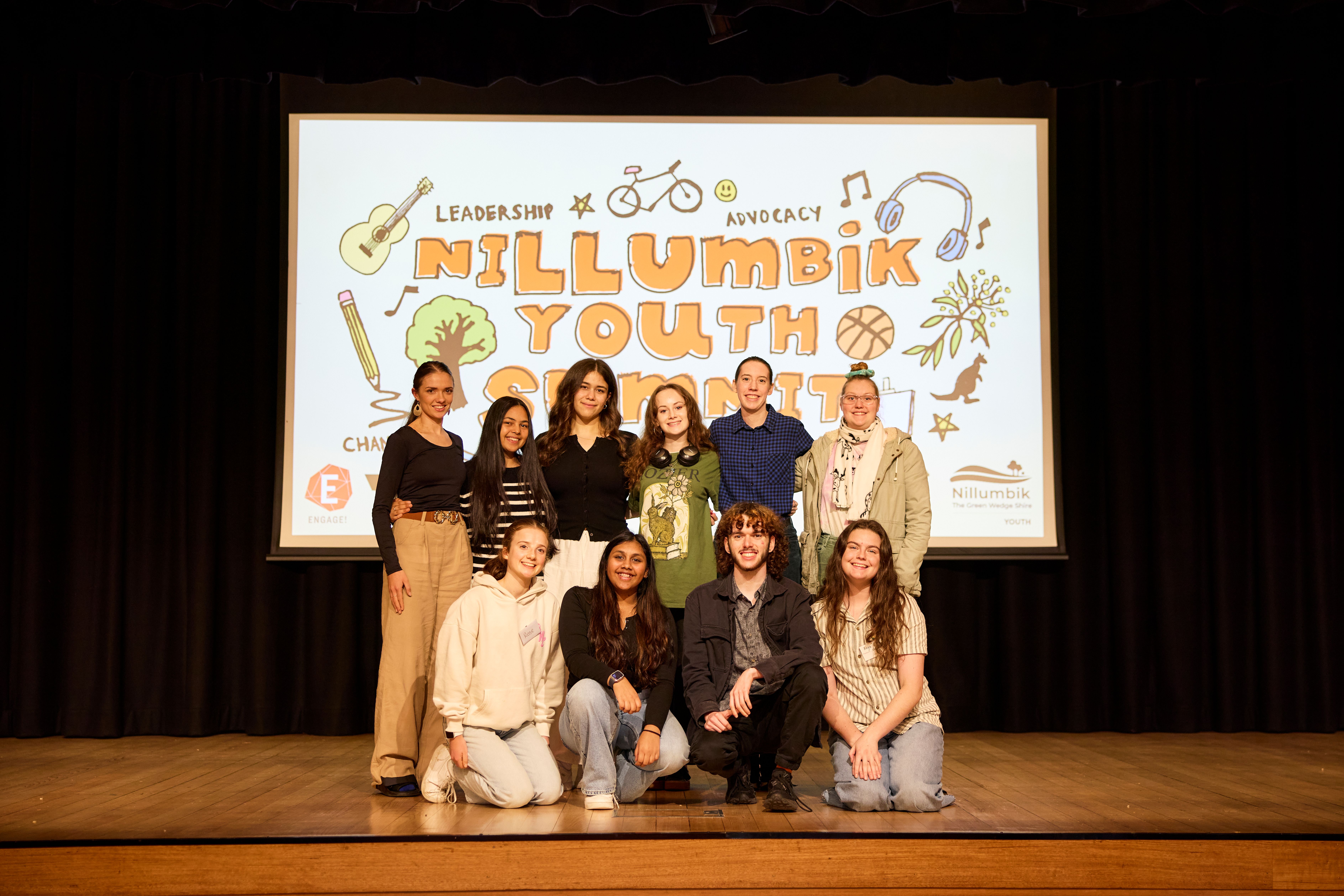 Ten members of Nillumbik Youth Council stand on a stage in front of a screen that says 'Nillumbik Youth Summit' on it. 
