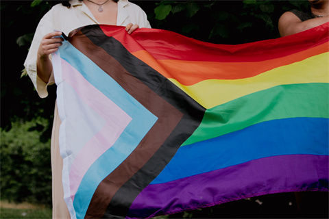Two people holding a large progressive pride flag in front of them. Image is cropped so you cannot see the people's faces.